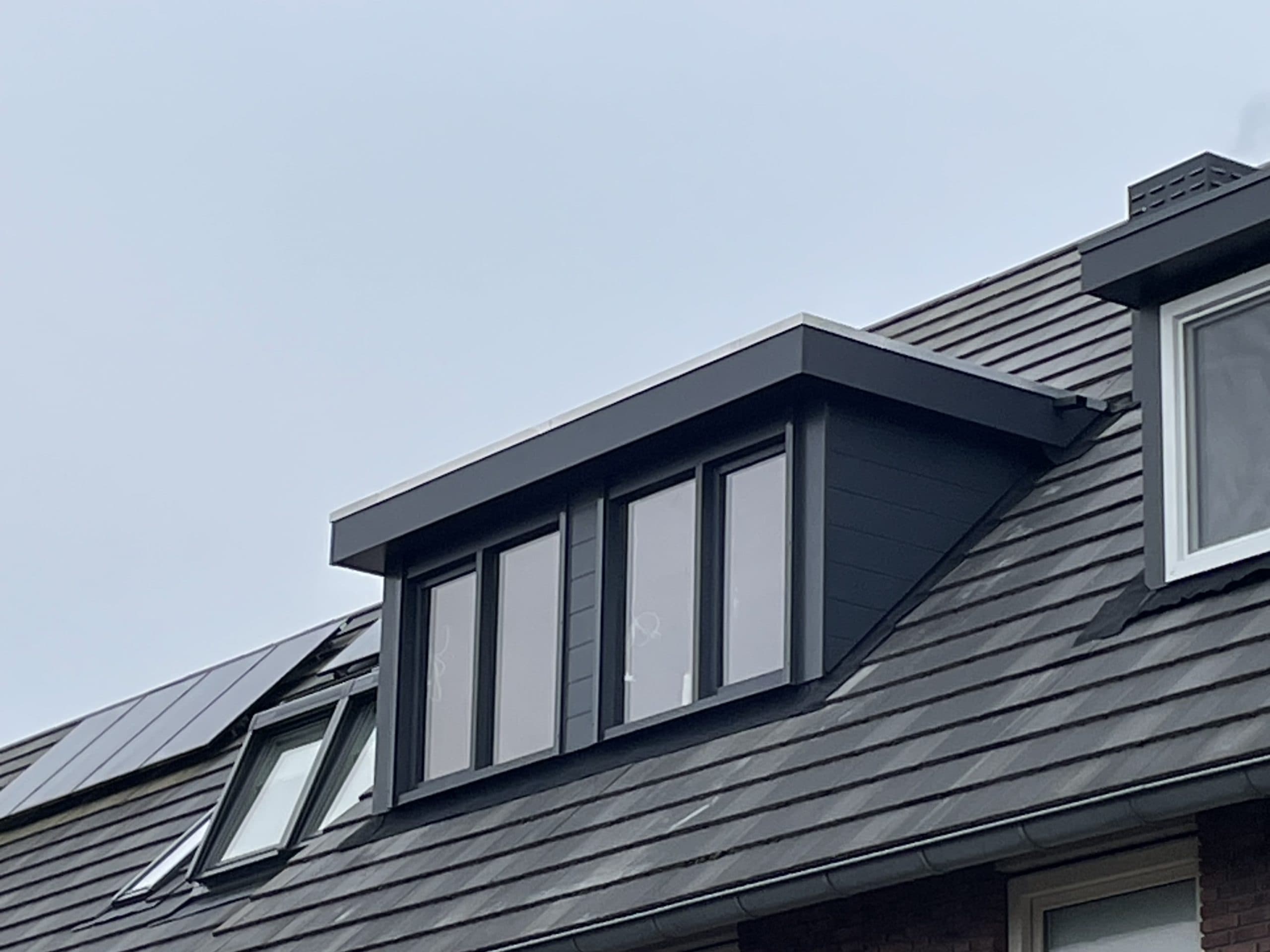 Een modern dak met een dakkapel, donkergrijze dakpannen en zonnepanelen aan de linkerkant, onder een bewolkte hemel.