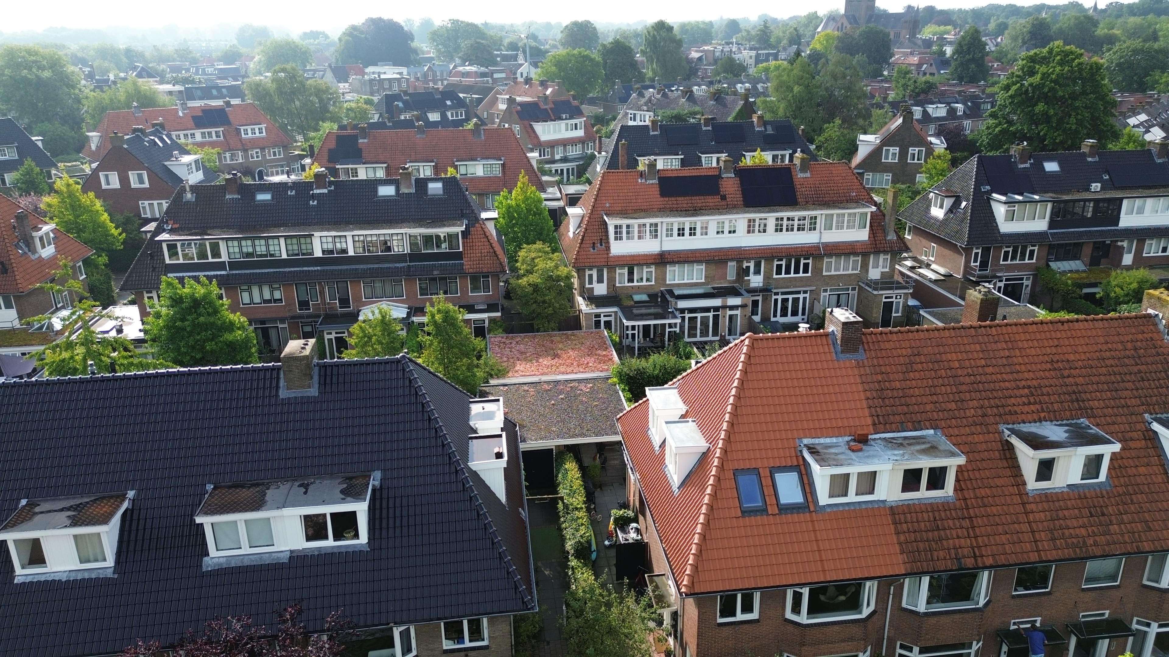 Luchtfoto van een buitenwijk met rijen gelijksoortige bakstenen huizen, rode en donkere daken en weelderige groene bomen op de achtergrond.